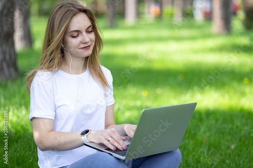 Female student in white t-shirt and jeans sit on the grass in spring park with notebook on her knees and prepare exams at University. Studying outdoors. Typing text on pc computer and listening music.
