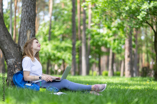 Modern young blonde girl in white t-shirt and jeans sit on the grass under tree with laptop on her legs and listen music with earphones. She smile and looks on trees tops. Coffee, notebooks, backpack