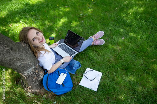 Freelance business concept. Top view of young woman who sits on the grass under tree with laptop on her legs. She look at camera and smile. Notebooks, coffee, dark-blue backpack, phone near.