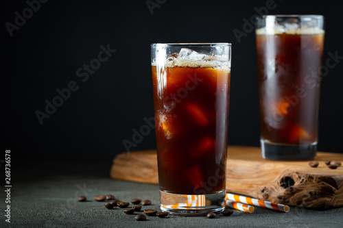Stampa su tela Ice coffee in a tall glass with cream poured over, ice cubes and beans on a old rustic wooden table