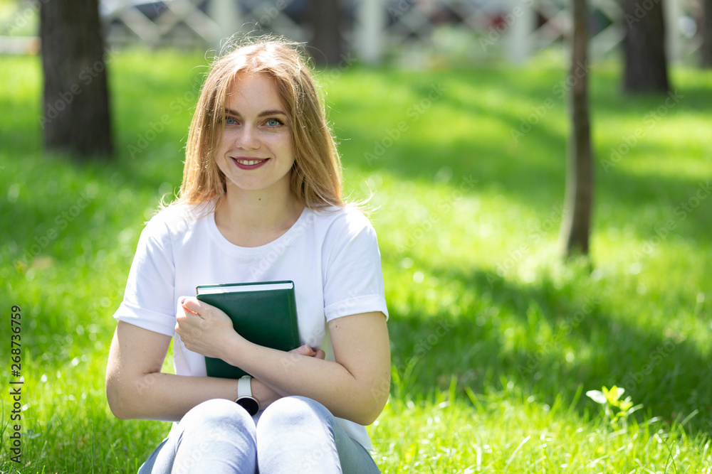 Smiling blonde girl-student in white t-shirt and jeans sits on the grass in sunny spring park and hug the book. Student studying outdoors. Space for text Smiling blonde girl-student in white t-shirt and jeans sits on the grass in sunny spring park and hug the book. Student studying outdoors. Space for text