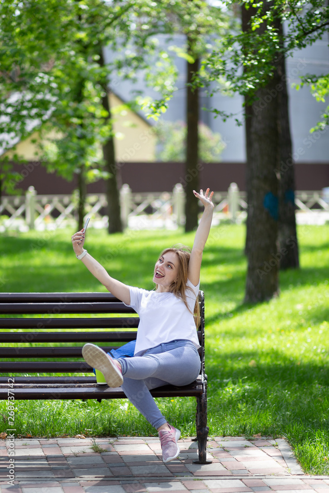 Very happy young blonde woman in white t-shirt and blue jeans sits on the bench with smartphone in her hand and makes selfie. Student lifestyle Very happy young blonde woman in white t-shirt and blue jeans sits on the bench with smartphone in her hand and makes selfie. Student lifestyle
