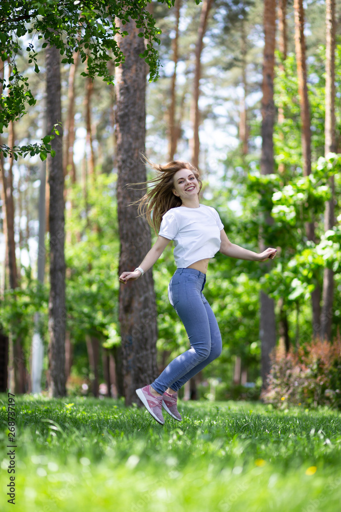 Naklejka premium Happy laughing blonde girl with white t-shirt, blue jeans and rose sneakers is jumping up on the green grass at the summer park.