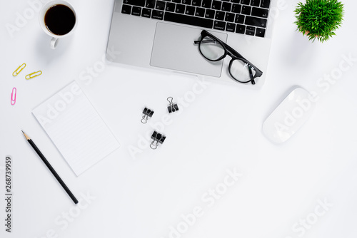The office desk flat lay view with laptop, mouse, tree, pink and yellow paper clip, coffee cup, notebook, pencil, black glasses on white background.