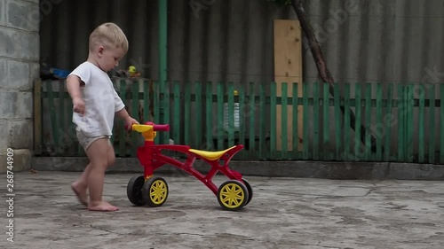 A blonde-haired toddler plays with a small bicycle in the yard of a private home