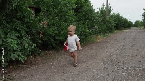 A blond-haired toddler with a baby red bucket runs down a path in the countryside