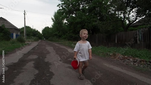 A blond-haired toddler with a baby red bucket walks down the road in the village