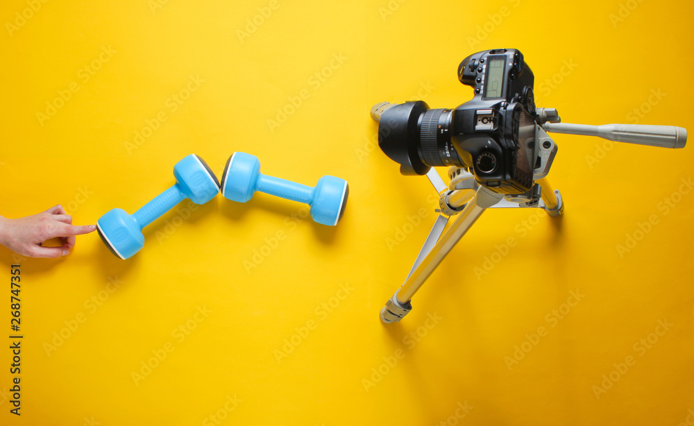 Fitness blogger. Woman Holding plastic dumbbells with her hands and blogging with camera on tripod on yellow background. Top view Fitness blogger. Woman Holding plastic dumbbells with her hands and blogging with camera on tripod on yellow background. Top view