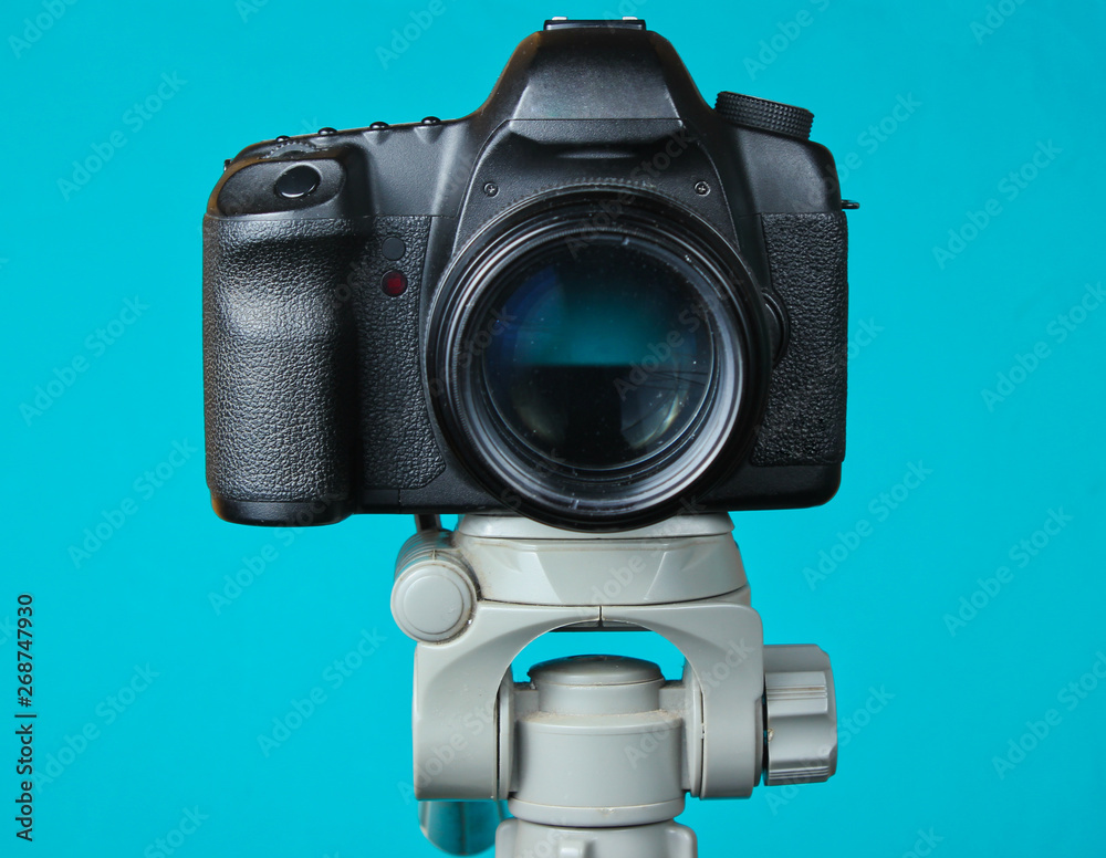 Modern digital camera with a tripod on blue background. Front view