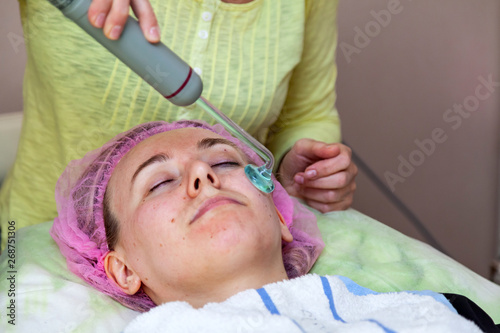 A young girl is lying on couch with a pink hat on her hair while performing an darsonvalization on face with a special device while cleaning and removing body fat under the skin on nose. Cosmetology photo
