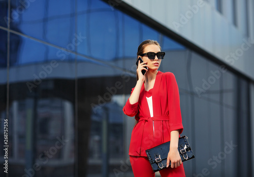 high fashion outdoor Portrait of a young woman in a red pantsuit talking on a mobile phone. Stylish black sunglasses, black clutch, long hair. © dinaphoto