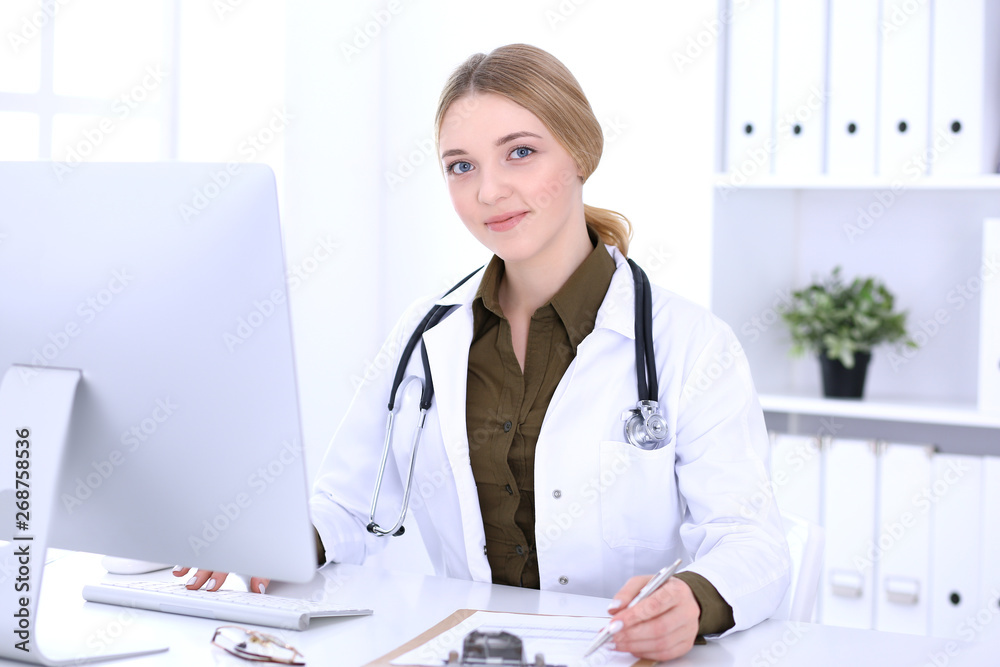Young woman doctor at work in hospital looking at desktop pc monitor. Physician controls medication history records and exam results. Medicine and healthcare concept