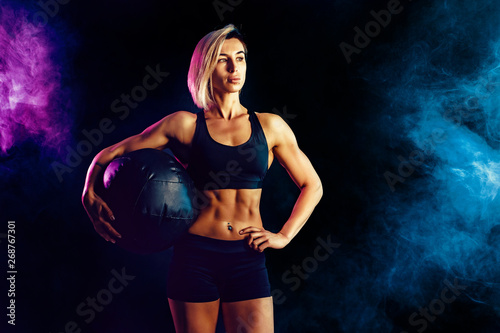 Sporty blonde woman in fashionable sportswear posing with medicine ball. Photo of muscular woman on dark background with smoke. Strength and motivation.