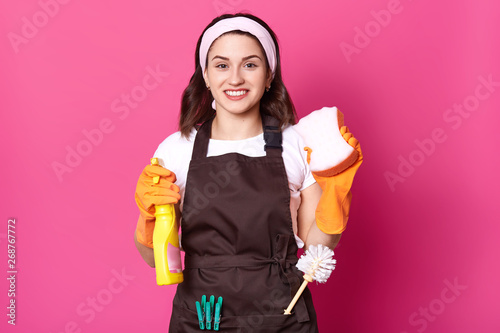 Cheerful positive, cute housewife holding washcloth and detergent in both hands, having toilet brush and clothespins in brown apron, wearing casual white t shirt and headband, looks delighted. photo