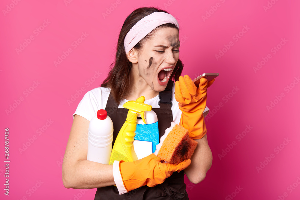 Studio shot of woman with cleaning materials holding phone in hand, looking on screen and screaming, has astonished and angry facial expression, has unpleasant conversation, isolated over pink wall. Studio shot of woman with cleaning materials holding phone in hand, looking on screen and screaming, has astonished and angry facial expression, has unpleasant conversation, isolated over pink wall.