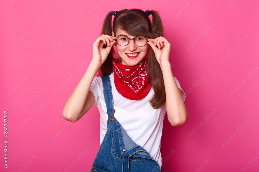 Image of joyful teenager girl wears round glasses, expresses positiveness,  dressed in fashionable clothes for youngsters, keeps hands on her eyewear,  isolated on pink wallPeople and happiness concept. Stock Photo | Adobe