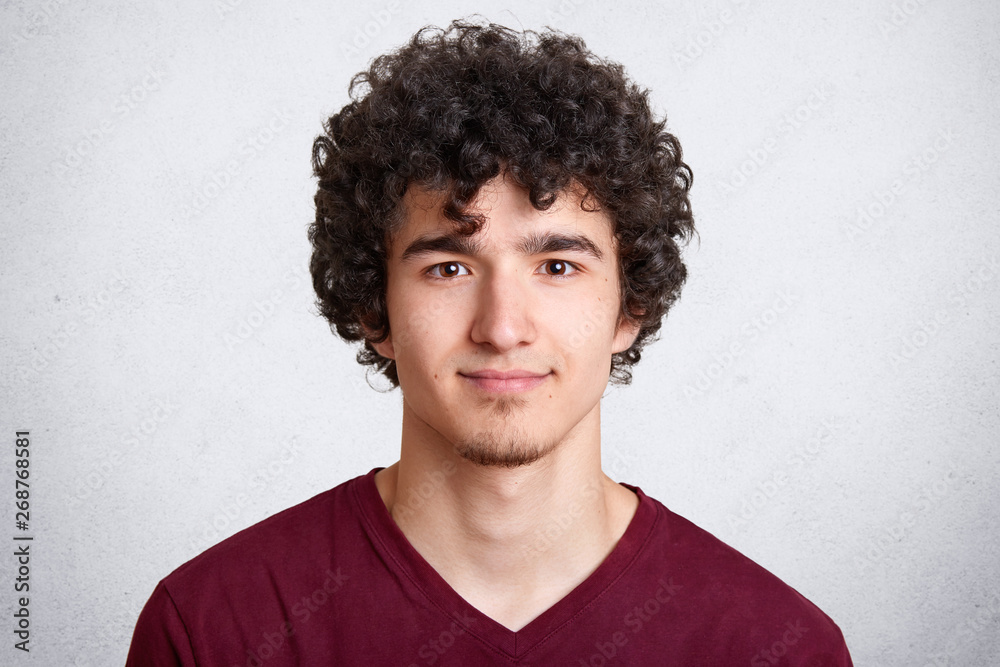 Obraz premium Close up portrait of curly handsome European male, wearing casual maroon shirt, isolated over white background, looking directly at camera, beig photographed in photo studio. People concept.