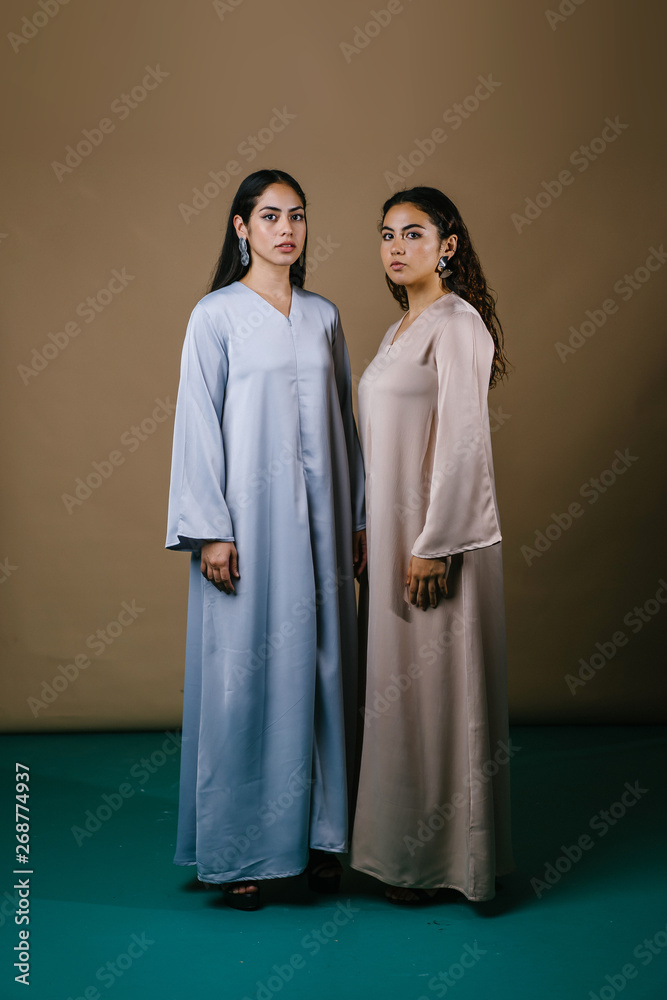 Portrait of two young Middle Eastern Muslim women as they pose for their photo. They are wearing traditional pastel coloured Baju Kurung dresses to visit during Raya and both have neutral expressions. Portrait of two young Middle Eastern Muslim women as they pose for their photo. They are wearing traditional pastel coloured Baju Kurung dresses to visit during Raya and both have neutral expressions.