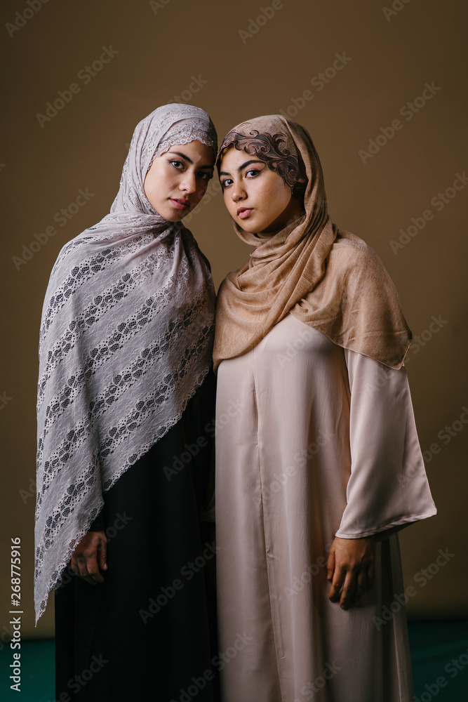 Two Muslim Middle Eastern sisters in traditional dresses and hijab head scarves stand and pose for a portrait together in a studio. They are both young, beautiful, elegant and attractive.