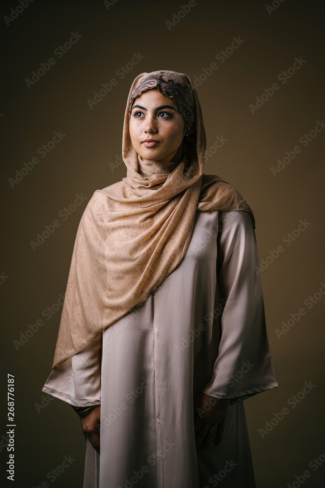 Portrait head shot of a young and beautiful Muslim Middle Eastern woman in a brown traditional outfit and hijab head scarf. She is stylish, elegant and fashionable. 