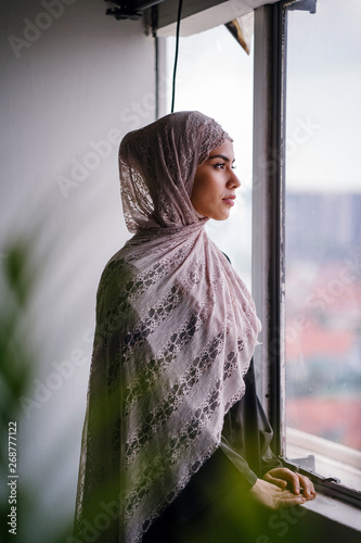 Portrait of a beautiful and tall Malay Asian Muslim woman in a hijab head scarf standing by the window of her home. She is wearing a traditional outfit and is about have begin her fast.  photo