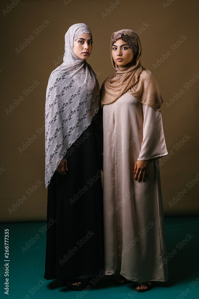 Two Muslim Middle Eastern sisters in traditional dresses and hijab head scarves stand next to another to pose for a portrait together in a studio. They are both dressed conservatively but stylishly. 