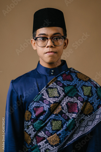 Studio portrait of a happy, handsome and young Malay Asian man in a blue baju melayu hat with a patterned sash dressed for Hari Raya. He is ready to go visiting friends and family during Raya.