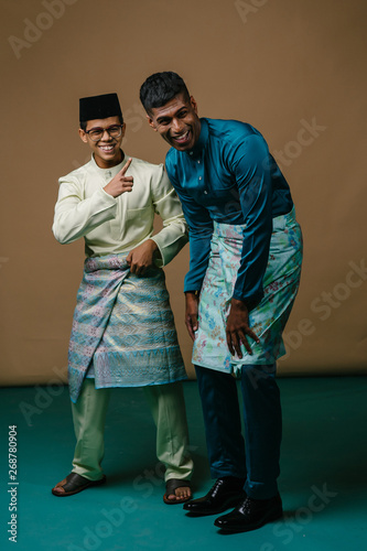 A young Malay and Indian men laugh and dance as they stand next to one another in a studio. They are both Muslims preparing to celebrate Ramadan and are both wearing traditional baju melayu clothing.  photo