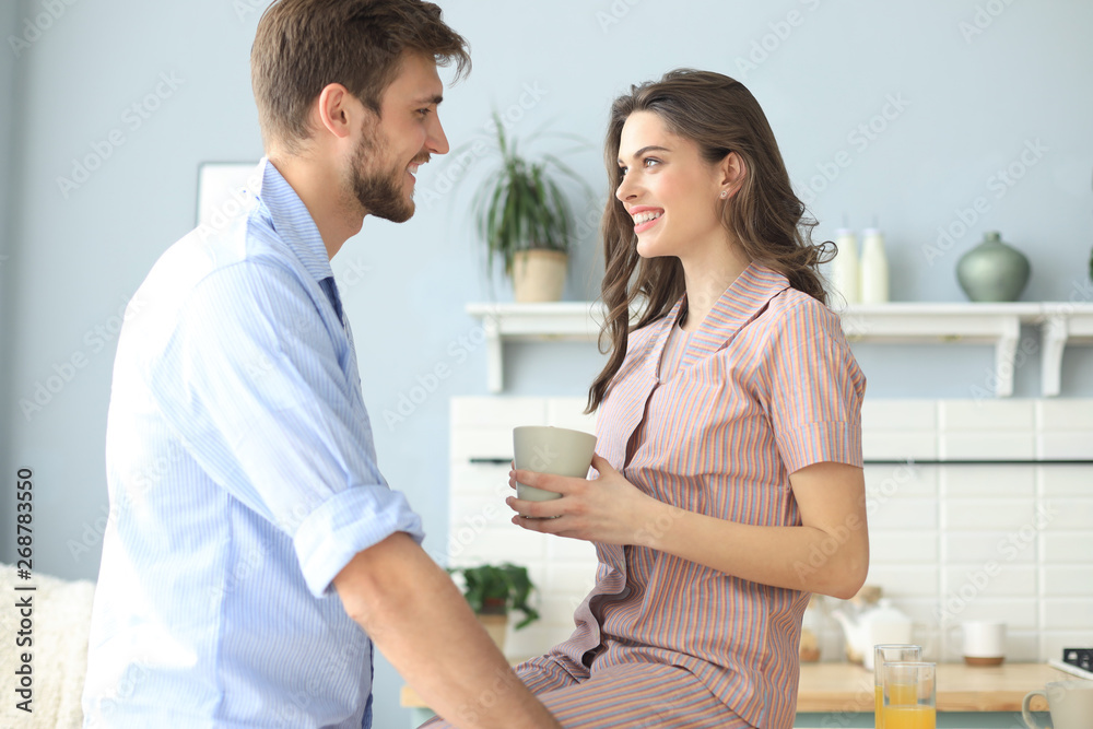 Fototapeta premium Beautiful young couple in pajamas is looking at each other and smiling while cooking in kitchen at home.