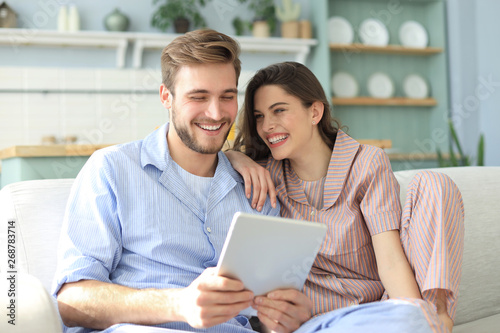 Young couple watching media content online in a tablet sitting on a sofa in the living room. © ty