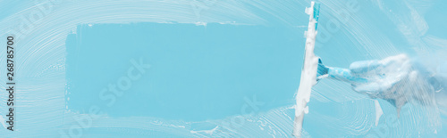 panoramic shot of woman in rubber glove cleaning glass with squeegee on blue background