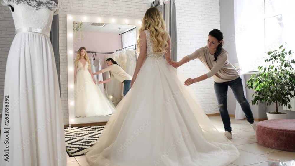 Wedding consultant helps bride choosen wedding dress in salon and wearing it near large mirror Wedding consultant helps bride choosen wedding dress in salon and wearing it near large mirror