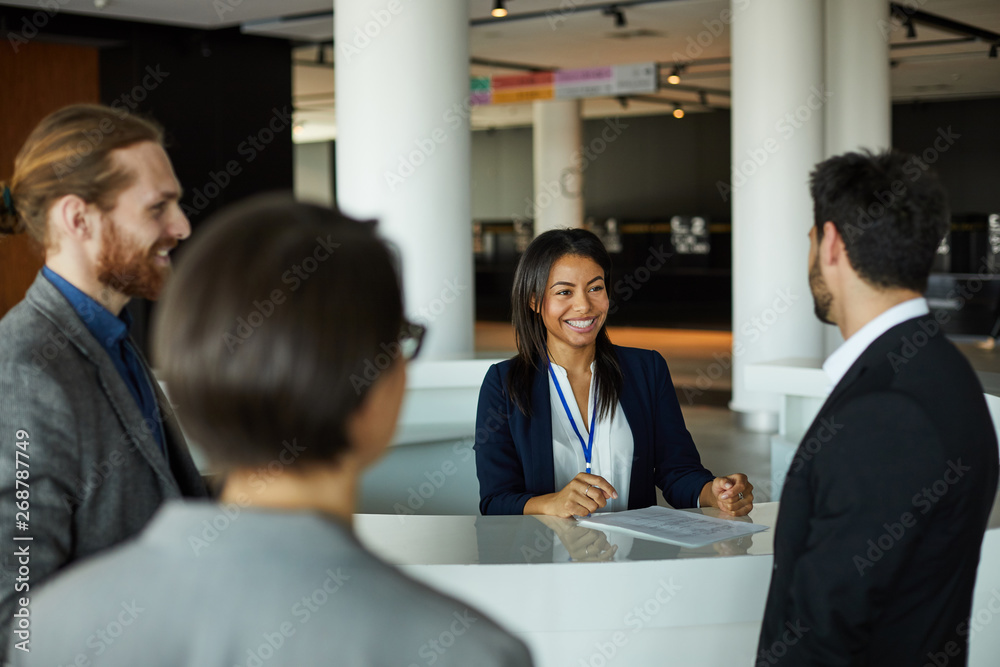 Jolly attractive young female administrator standing at counter and checking list while registering people for participation in forum Jolly attractive young female administrator standing at counter and checking list while registering people for participation in forum