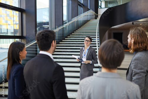 Smiling confident business forum guide with badge on neck standing on stairs and holding clipboards with files while giving tour to participants photo