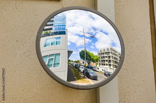 Fotografie Outdoor convex safety mirror hanging on wall with reflection of an urban roadside view of cars parked along the street by residential apartment buildings
