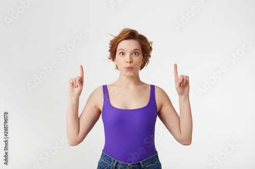 Portrait of a young happy amazed attractive short-haired girl, wearing a purple jersey, looking at the camera, speaks lips "wow" and index fingers point up at the copy space isolated over white wall.