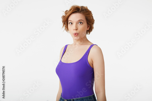 Photo of amazed young attractive short-haired girl, wearing a purple jersey, amazed looking up at the copy space, speaks lips "wow" isolated over white background.