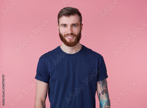 Portrait of young happy attractive red-bearded guy with blue eyes, wearing a blue t-shirt, smiling and looking at the camera isolated over pink background.