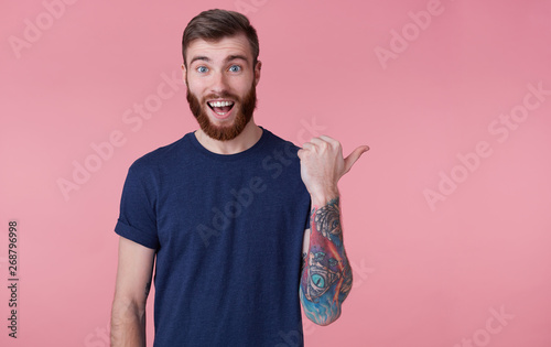 Prttrait of young happy red-bearded young guy ,with wide open mouth in surprise, wearing a blue t-shirt, pointing finger to copy space at the right side isolated over pink background.