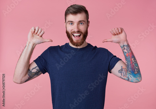 Portrait of young happy attractive red-bearded young guy , wearing a blue t-shirt, broadly smiling, enjoy himself, with arms raised up and pointing fingers at himself isolated over pink background.