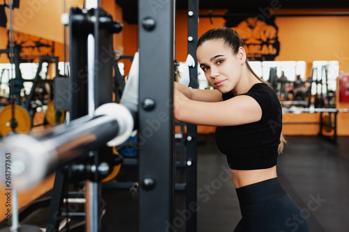Portrait of a beautiful young posing woman standing near the bar preparing for the exercise. Concept of strong-willed and trained professional athlete