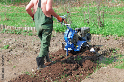 A farmer in rubber boots and overalls plowing the ground with a cultivator on a sunny day.