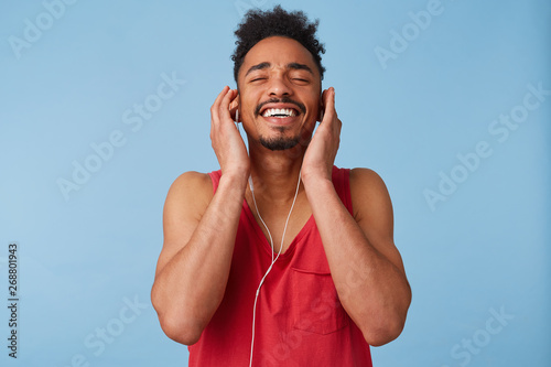 Фотография Young african american man feeling great and very happy, close eyes and enjoying, listening with headphones to favorite song, tilted his head up and broadly smiling isolated over blue background