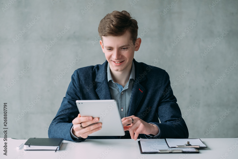 Successful career. Happy office worker. Young man sitting at desk, using tablet, smiling. Copy space. Successful career. Happy office worker. Young man sitting at desk, using tablet, smiling. Copy space.