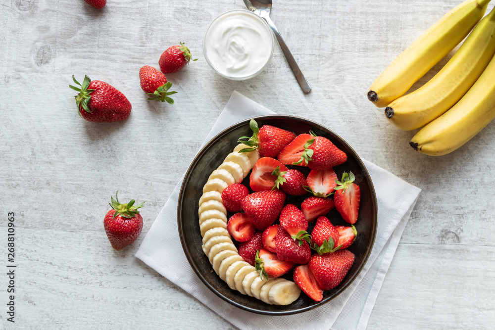 Salad of strawberries and banana on a dark plate on a light background. Served with Greek yogurt. Bananas and strawberries. Top view. Salad of strawberries and banana on a dark plate on a light background. Served with Greek yogurt. Bananas and strawberries. Top view.