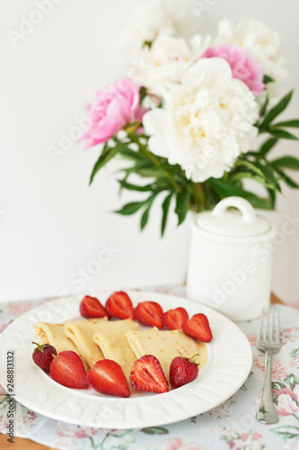 pancakes with strawberries and coffee on the table near a vase with peonies on a white background