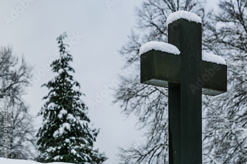 Winter scene of Christian stony cross at a cemetery and trees in the background covered with snow.