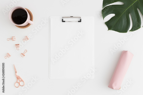 Top view of a white clipboard mockup with  workspace accessories, coffee and a monstera leaf on a white table.