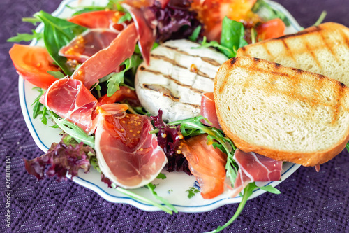 Tasty mixed salad dish with grilled camembert cheese, prosciutto ham, organic tomato and fresh green leaves. Healthy dilicious meal. Isolated on black