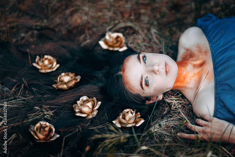 portrait of beautiful brown-haired woman in a blue dress lying on grass, with golden roses on her long hair, in the fairy forest, golden paint shimmer on her neck fantasy concept - obrazy, fototapety, plakaty 